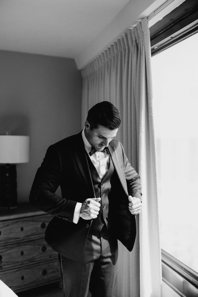 Black and white photo of a handsome groom buttoning up his jacket near the window of his hotel room, looking down. 