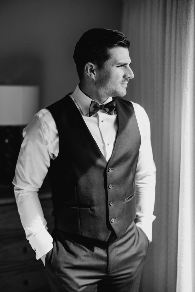 Black and white photo of a groom standing by the window ready for his bride to get ready.