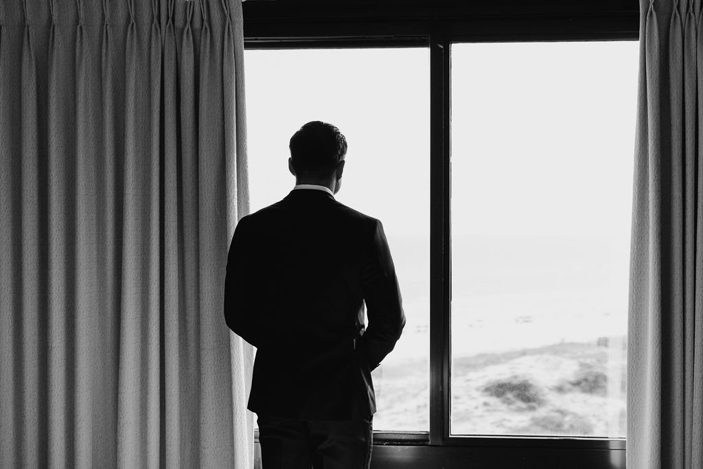 Black and white photo of a handsome groom taken from behind, while he's looking out in the distance through a window of his hotel room.